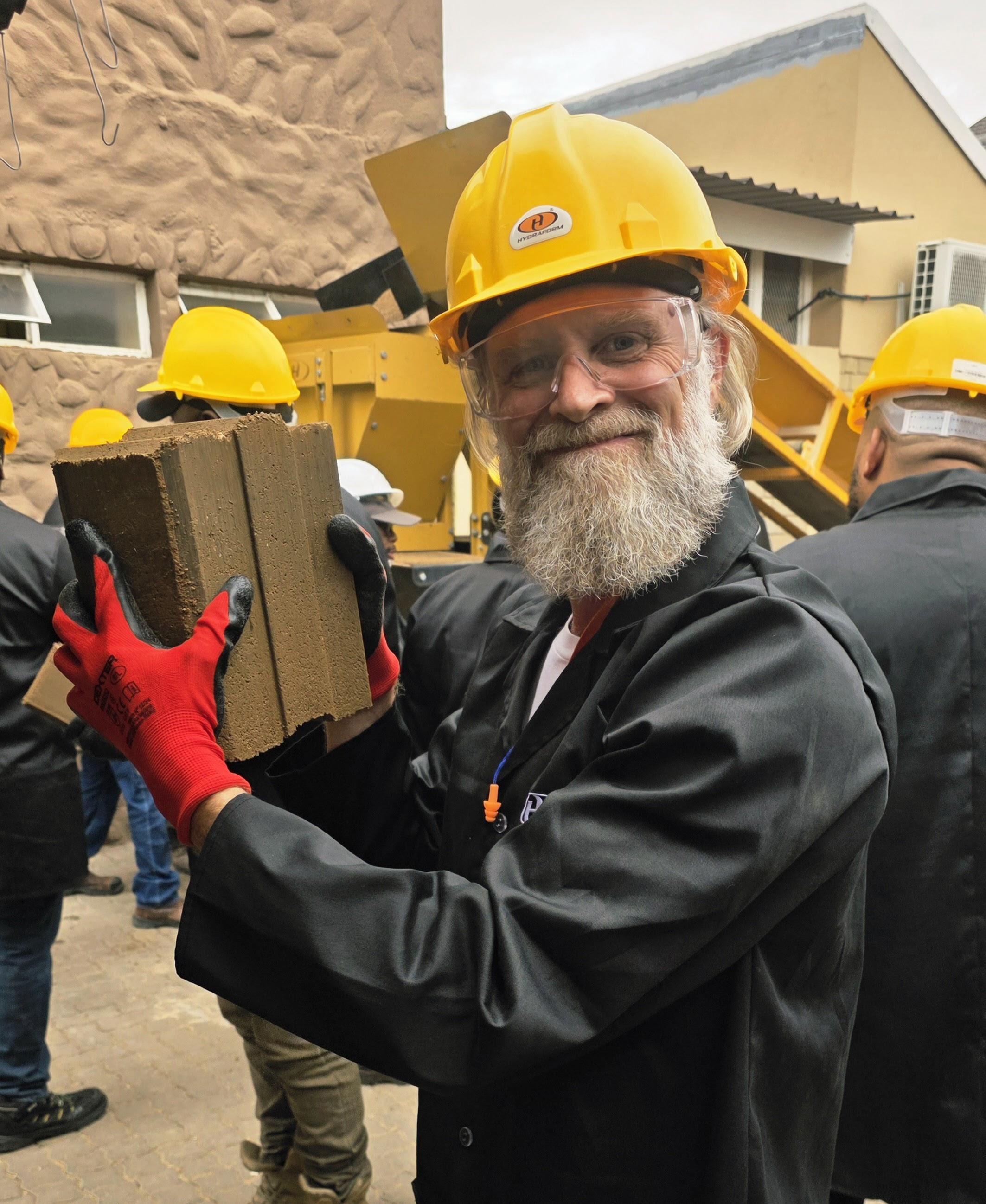 Kieran Shirey holding a freshly made compressed earth block beside production equipment
